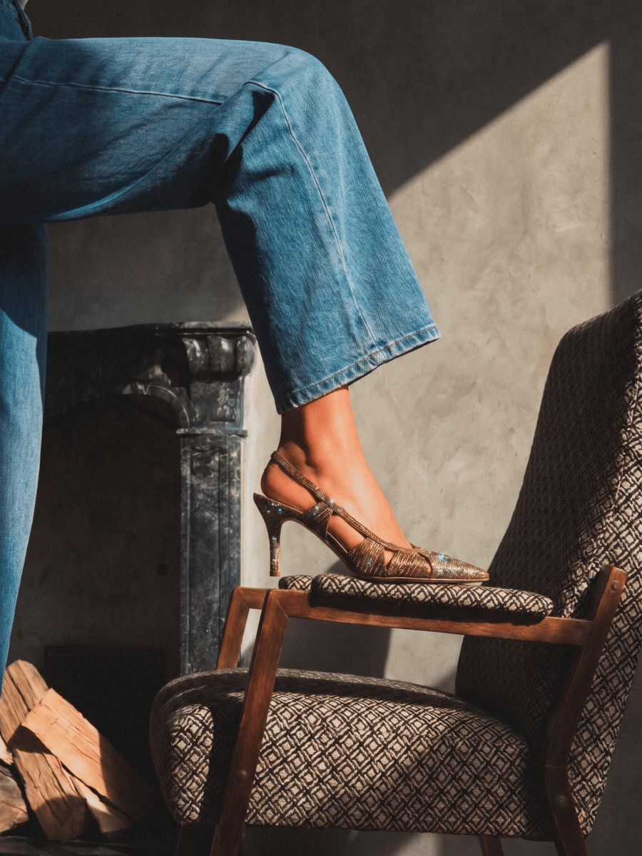 A person wearing blue jeans sits with one leg raised on the cushion of a patterned wooden armchair, wearing brown strappy slingback high heels in a sunlit room.
